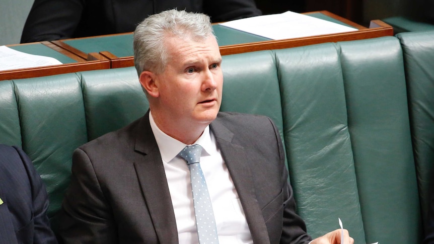 Burke with a confused expression, looks across the chamber, wearing a light blue tie and holding a piece of paper.