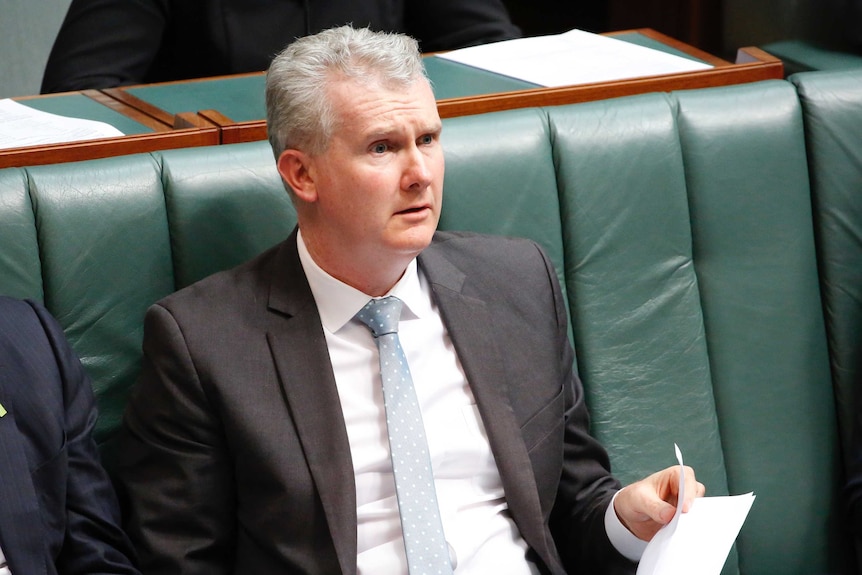 Burke with a confused expression, looks across the chamber, wearing a light blue tie and holding a piece of paper.