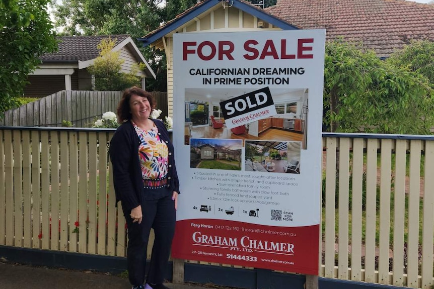 Sandra wears a bright flowery top and black cardigan standing in front of a for sale sign with a sold sticker