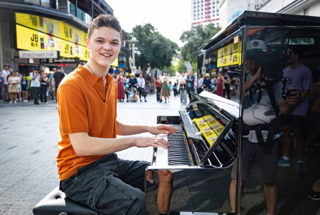 A smiling young man in an orange shirt is seated at a black upright piano in the middle of a busy pedestrian area.