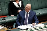 A bald man in a suit holds a pen as he sits behind a desk in parliament.