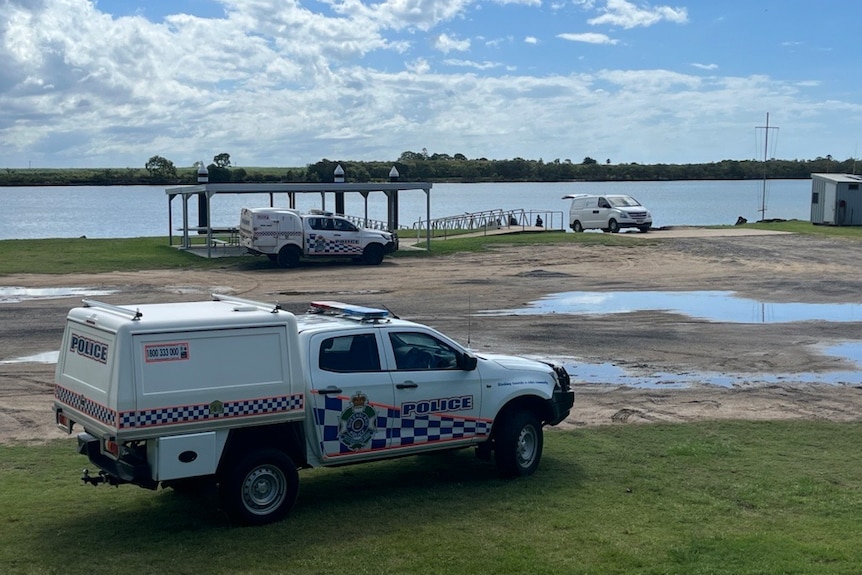 Three police cars by the side of the river, parked on gravel and grass in front of a jetty and a boat ramp.