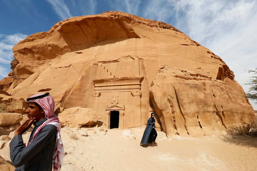 A woman with a camera walks in front of a stone tomb.