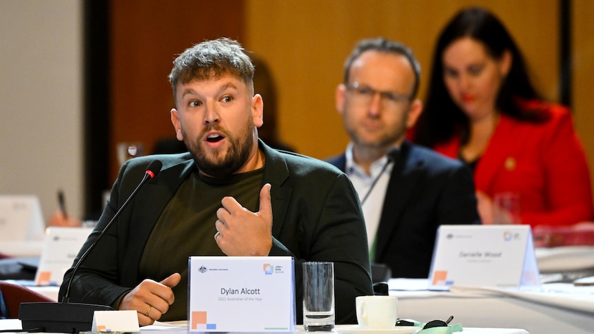 Dylan Alcott speaks into a microphone and gestures with his hand while sitting at a desk with his name printed on a sign.