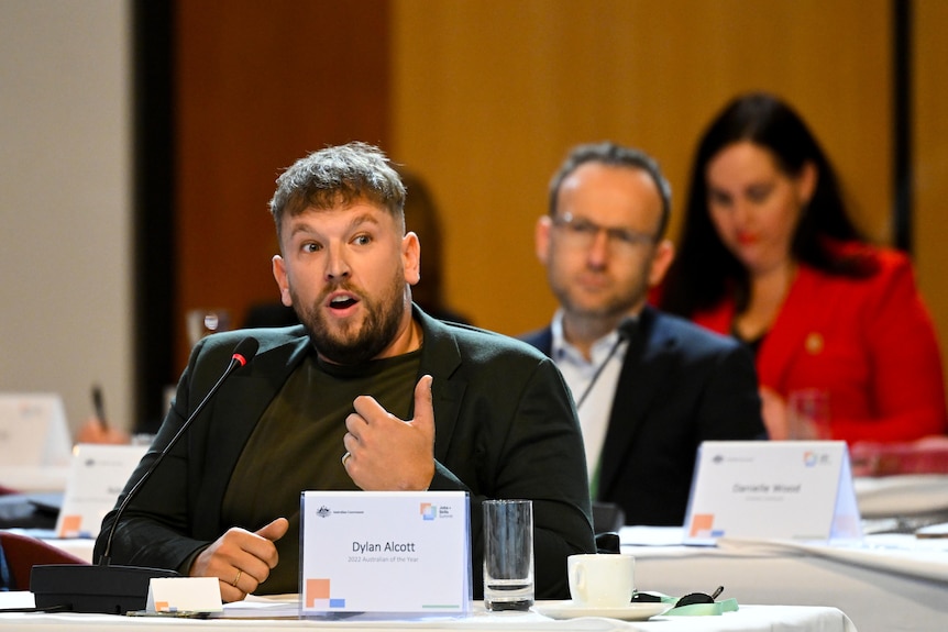 Dylan Alcott speaks into a microphone and gestures with his hand while sitting at a desk with his name printed on a sign.