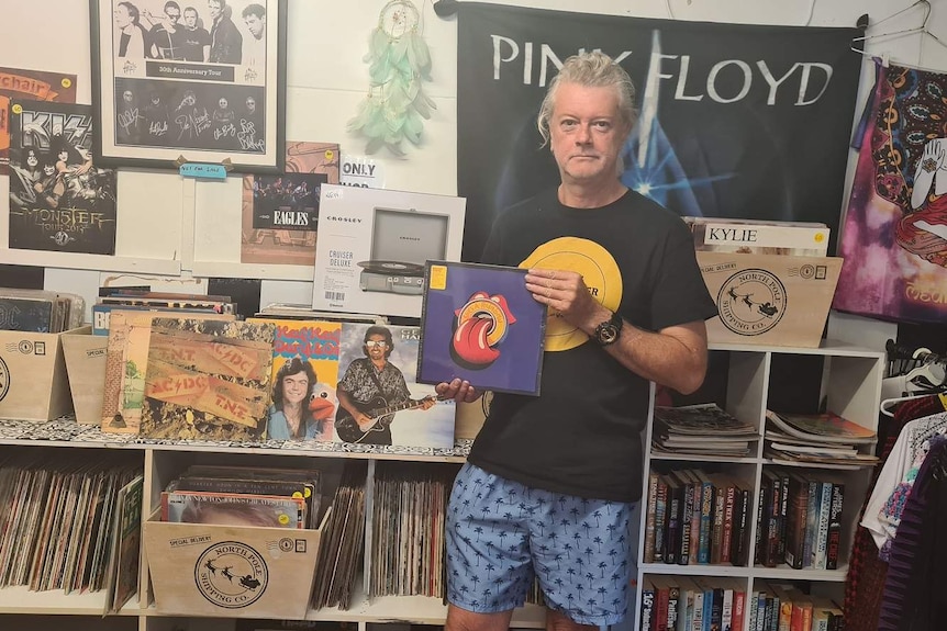 Man with long, grey hair stands in front of record collection and holds record in hands