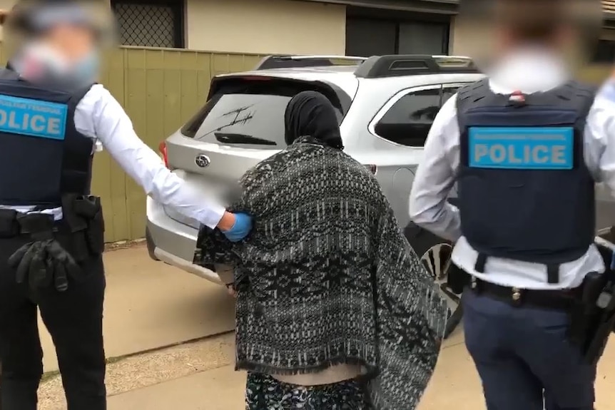 Two police officers wearing bulletproof vests stand either side of a woman wearing a face covering, walking to a police car.