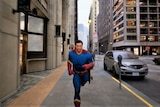 A caped superhero runs along a city street in a video game towards the camera