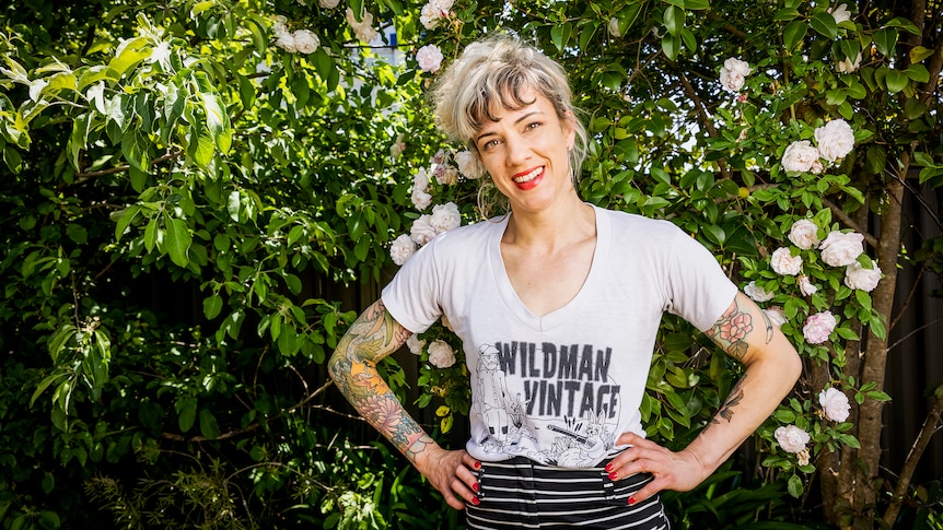 Jenny Valentish, smiling widely, stands with hands on hips with fair hair loose on hear and tshirt with text 'Wildman vintage'.