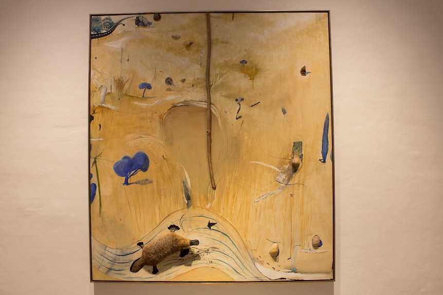 A painting of abstract yellowed paddocks with blue trees and a real branch and stuffed platypus
