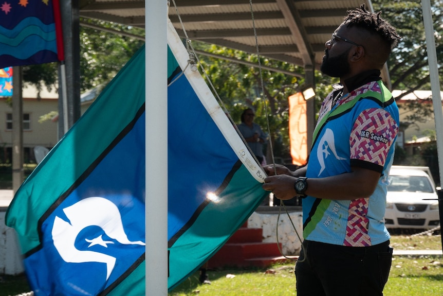 An indigenous man stands at a flag pole preparing to raisea blue, aqua and black striped flag with a 5-pointed white star.  