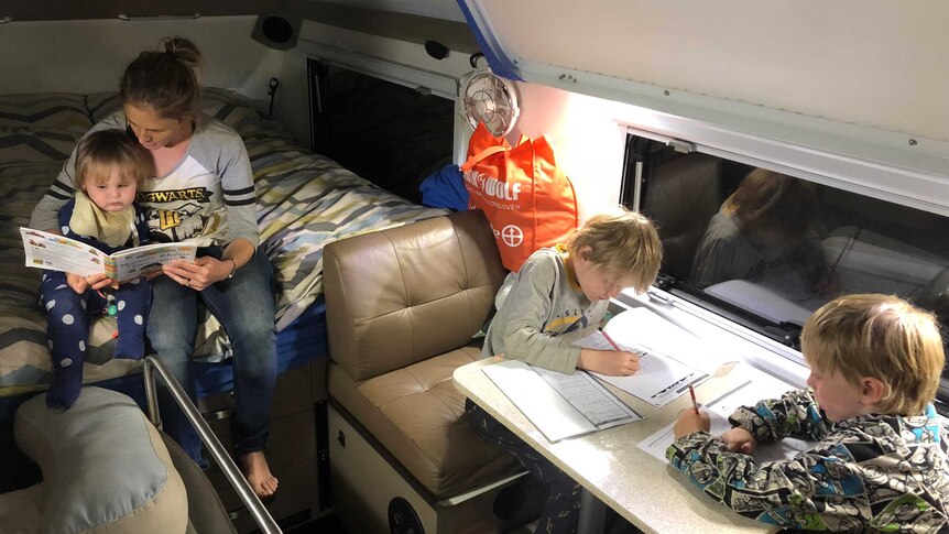 Amy Murphy reads to her baby son Wade, while Jake (9) and Lucas (8) do their school work at a small table inside their camper.