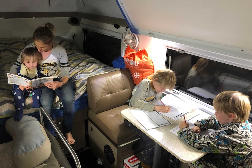 Amy Murphy reads to her baby son Wade, while Jake (9) and Lucas (8) do their school work at a small table inside their camper.