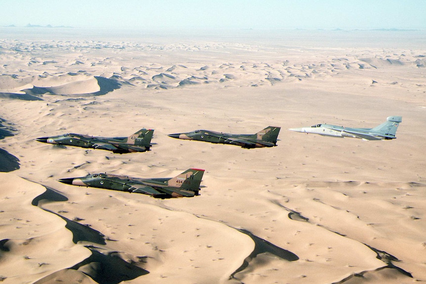 An aerial photo shows four fighter aircraft over desert sands, three of which are in green camouflage.