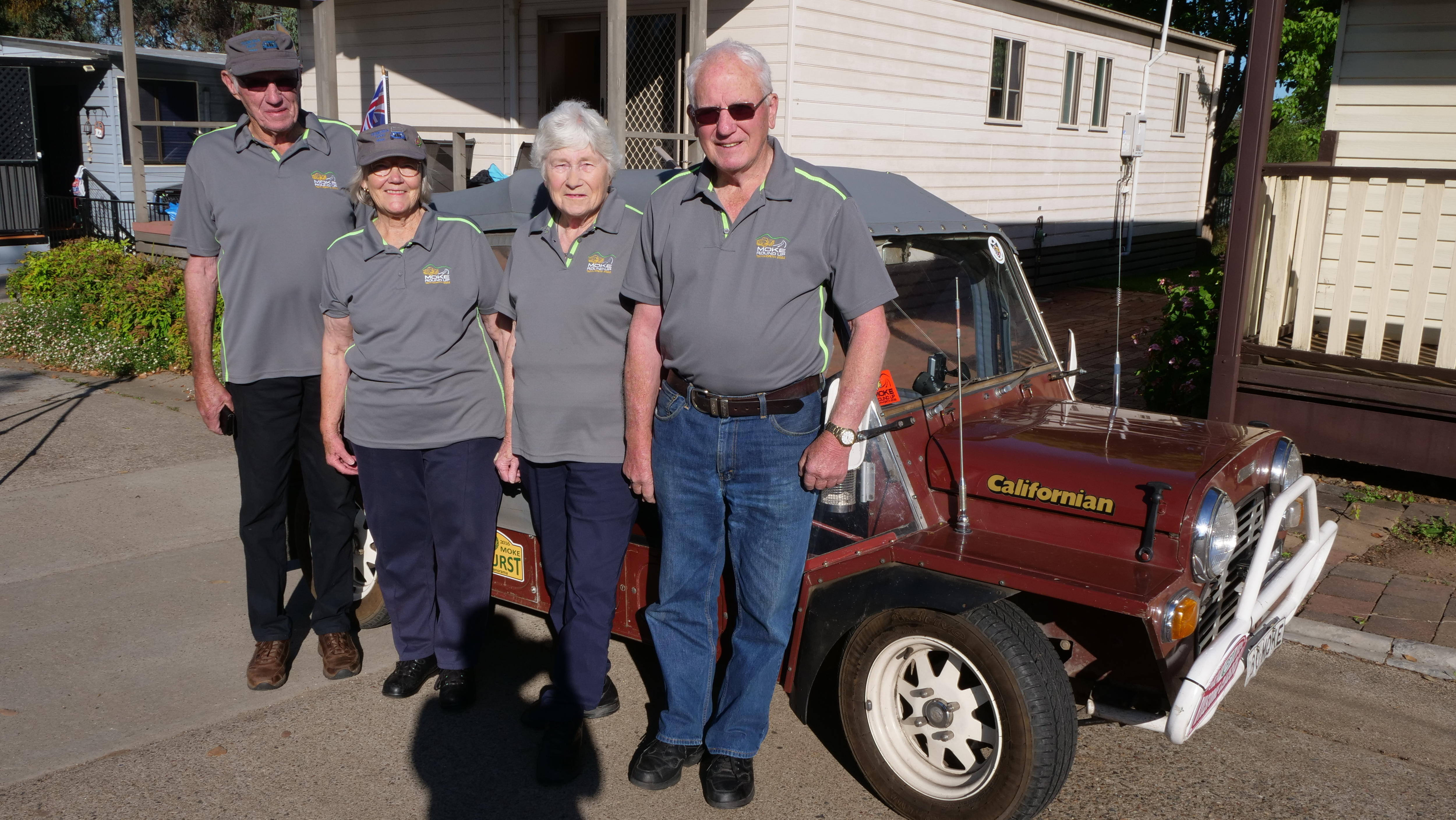 Two men and two women in matching grey shirts stand in front of a maroon Mini Moke.