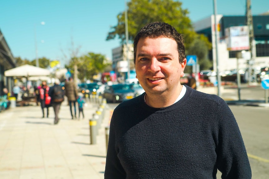 A man in a white t-shirt and navy jumper smiling on a street corner