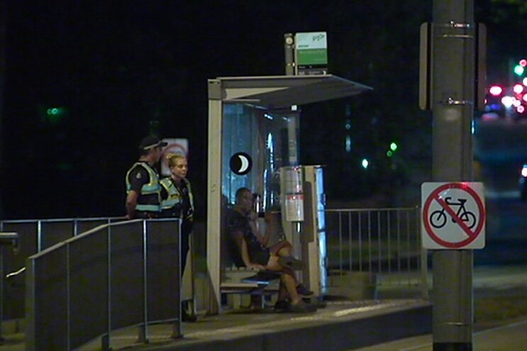 Uniformed police officers in fluorescent high-visibility jackets stand at a tram stop at night.