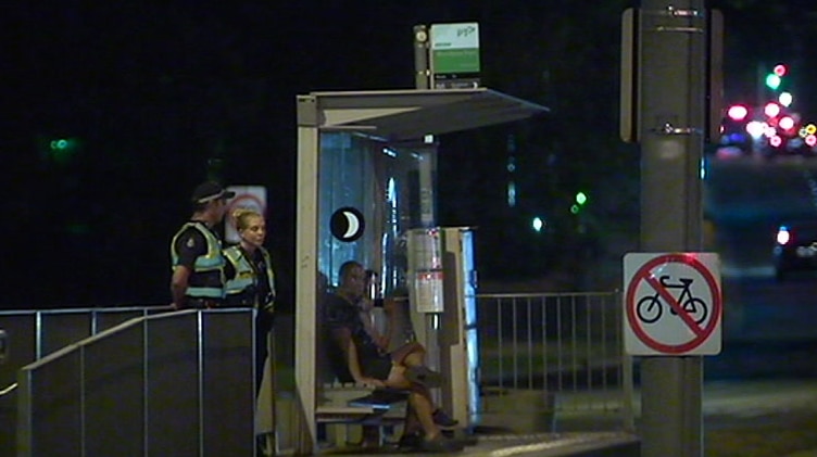 Uniformed police officers in fluorescent high-visibility jackets stand at a tram stop at night.