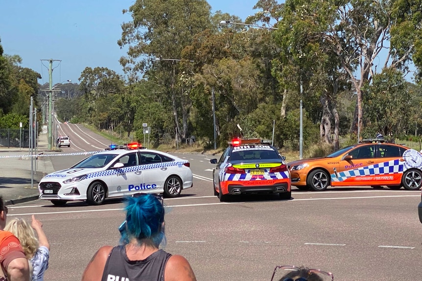 Three police cars and police tape block off a road while onlookers stand in the foreground looking.