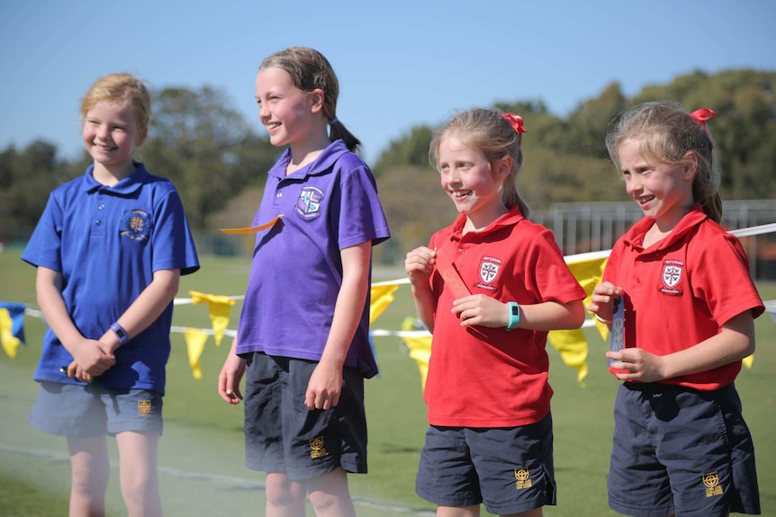 Four girls stand with their winning ribbons on an oval after a running race at a sports carnival.