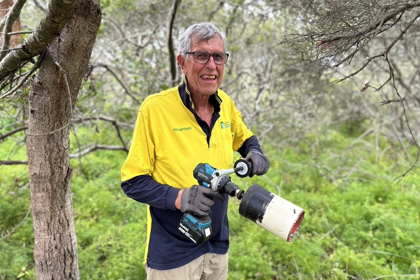 A man with grey hair wearing glasses and a yellow polo shirt holds a tool resembling a drill.