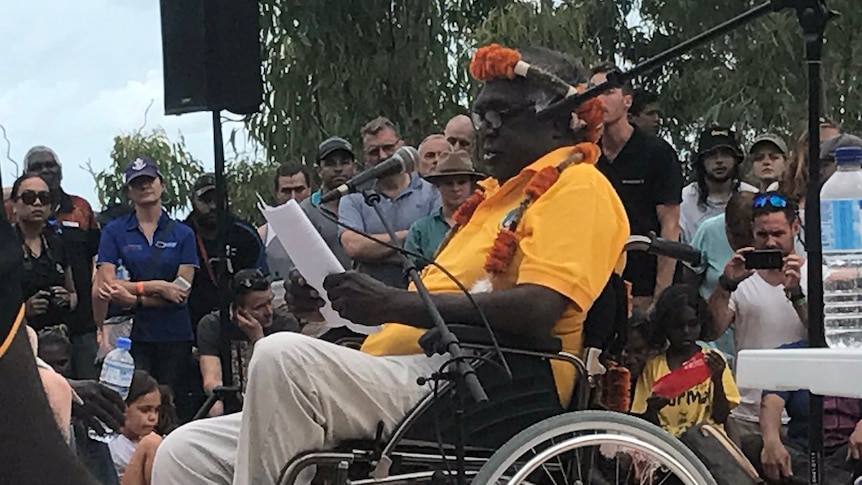 Galarrwuy Yunupingu sits in a wheelchair at a microphone and reads from a piece of paper.