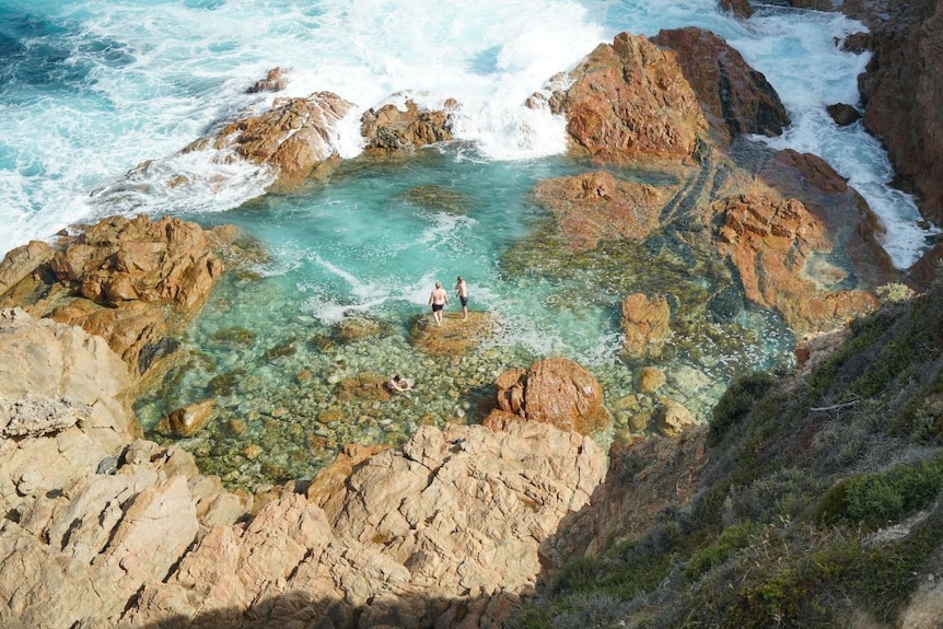 Two men stand on a rock next to a the ocean and surrounded by boulders