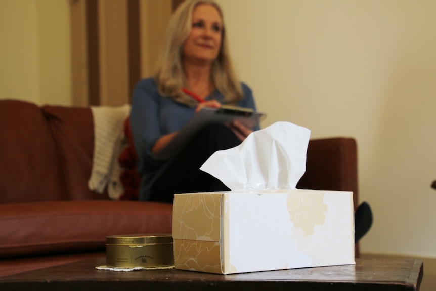 A tissue box sits in the foreground on a coffee table, with a blurred image of a woman with blonde hair sitting on a couch.