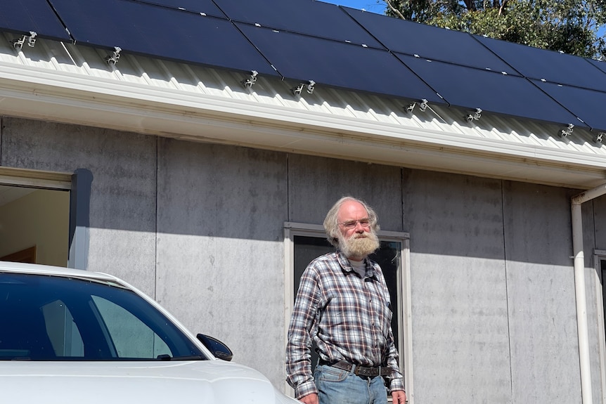 Eric stands in jeans and a pleated shirt to the right of his electric vehicle and in front of his home.