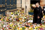 Charles and Camilla look at piles upon piles of flowers outside a grand black and gold gate