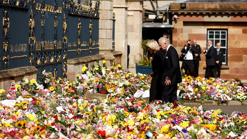 Charles and Camilla look at piles upon piles of flowers outside a grand black and gold gate