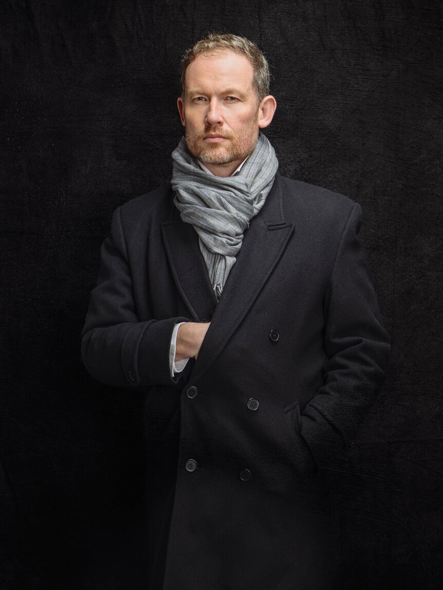 Photographer Charles Brooks stands in a dark coat with the light blue scarf. One hand sits inside his coat.