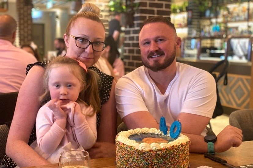 A woman holds a young girl as they sit at a table with a man in front of a 40th birthday cake.