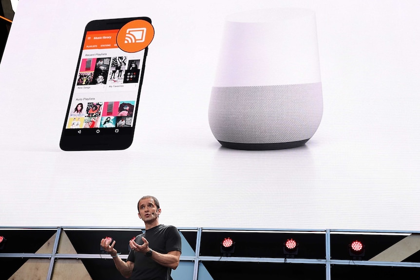 A man talks in front of a large screen showing a mobile phone and a Google Home device.