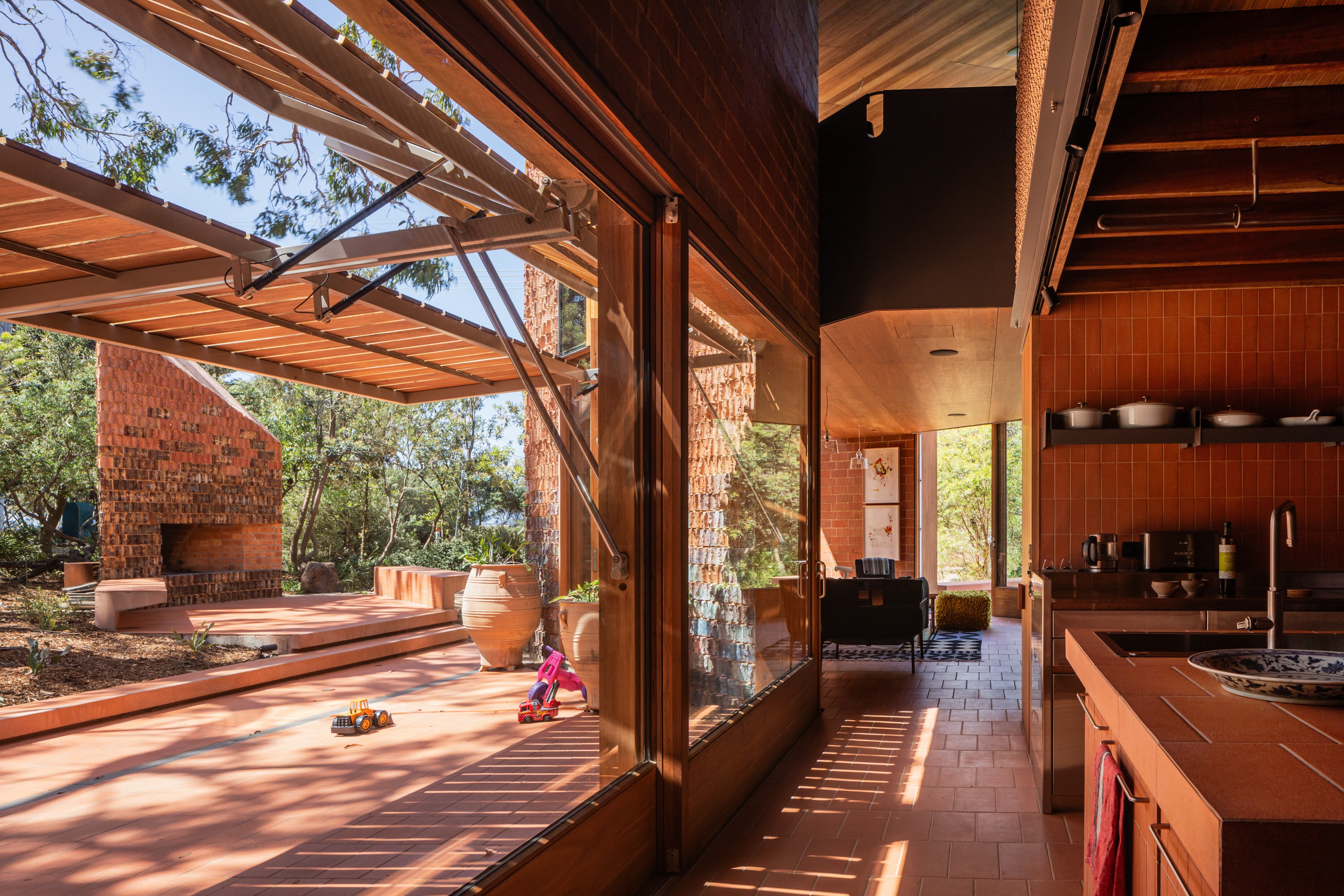 Wide open windows overlook green trees, with an angular house constructed of red brick and rich wood