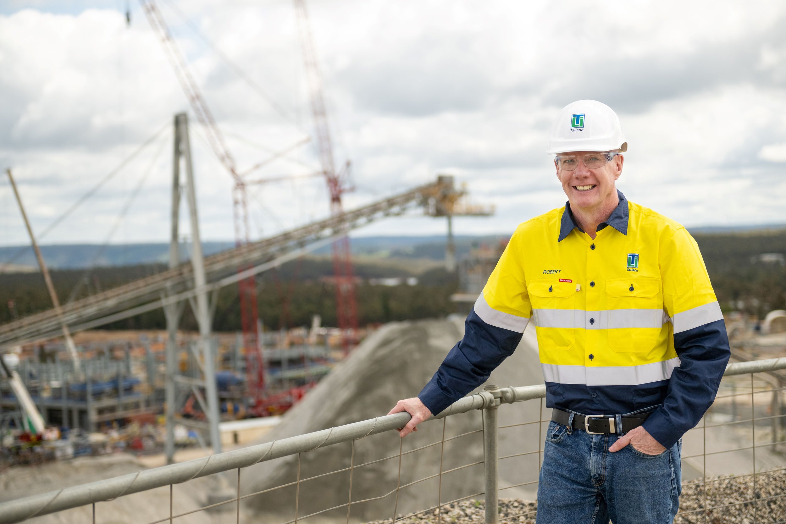 Un hombre con un chaleco reflectante y un casco protector se para frente a la empresa minera y mira a la cámara, sonriendo