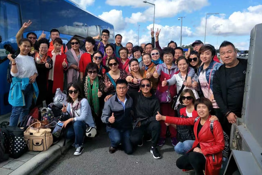 An Aus Highway Travel Services tour group poses for a photo next to a bus making thumbs up and peace signs.