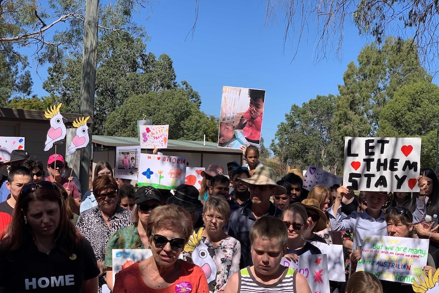 A small crowd of people, including children, hold signs saying 'let them stay' and pictures.