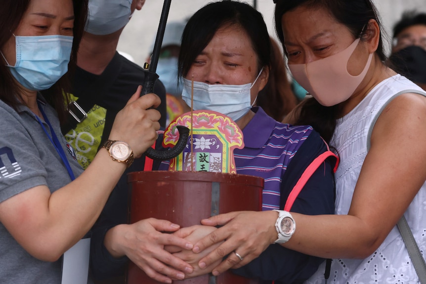 A weeping woman is comforted by two other women while holding a large brown pot. 