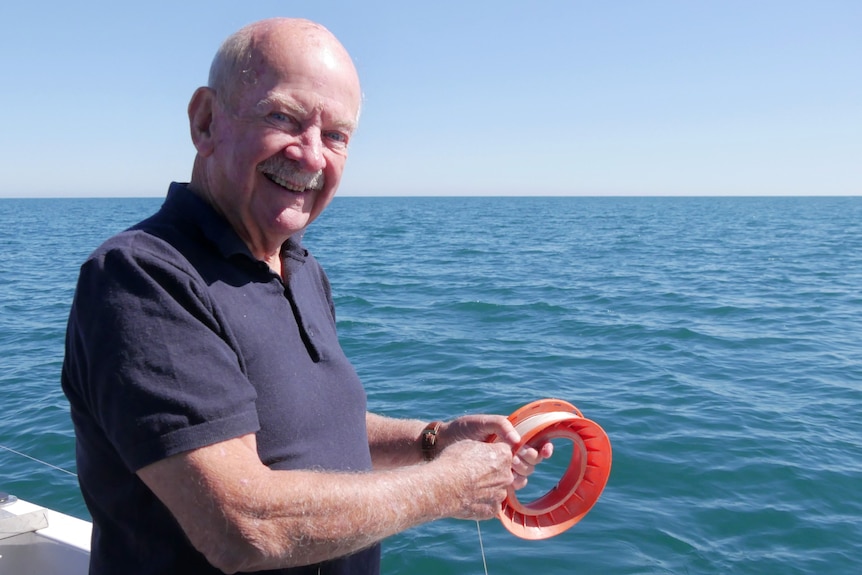 A many in a navy polo shirt smiles at the camera while holding a handheld fishing reel, with a line dropped into the sea.