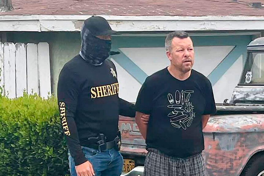 a man in a face mask and black sheriff shirt next to a man with his hands behind his back