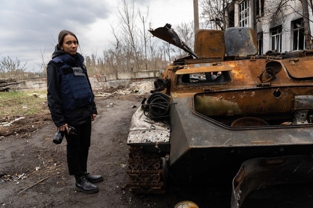 Woman in bullet-proof vest, holding a camera standing next to a burnt out tank.