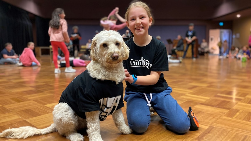 A young girl and a fluffy dog sit on ground of rehearsal space with cast behind