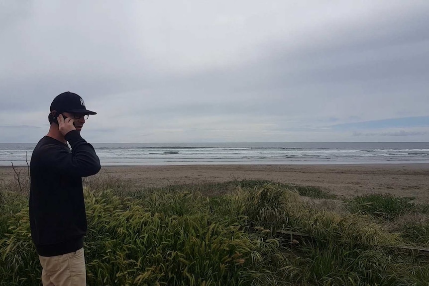 A man in a black sweater and baseball cap making a phone call at the beach.