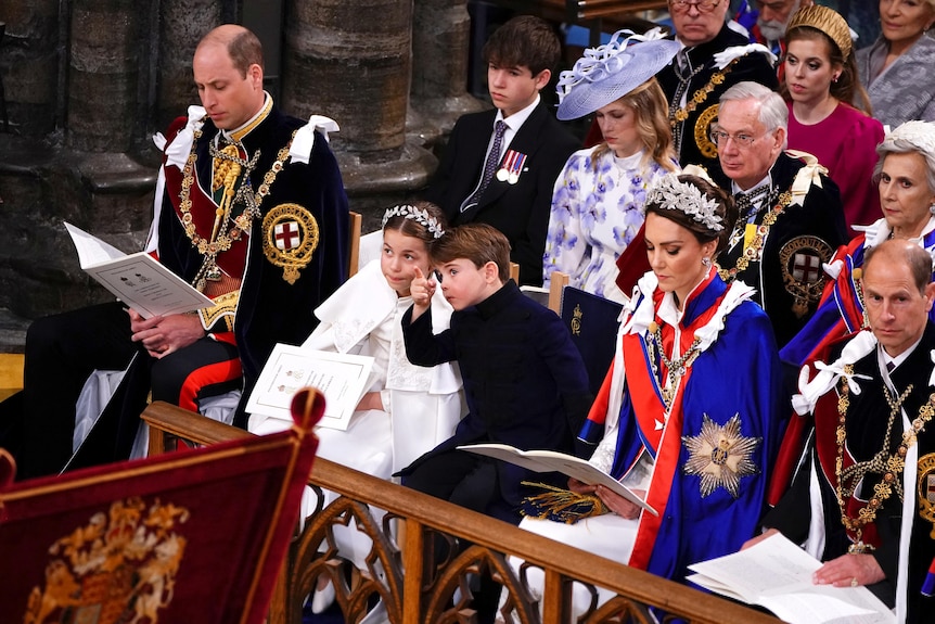 Charlotte and Louis whisper and point in the Abbey. Seated either side of them, Wills and Kate read programs