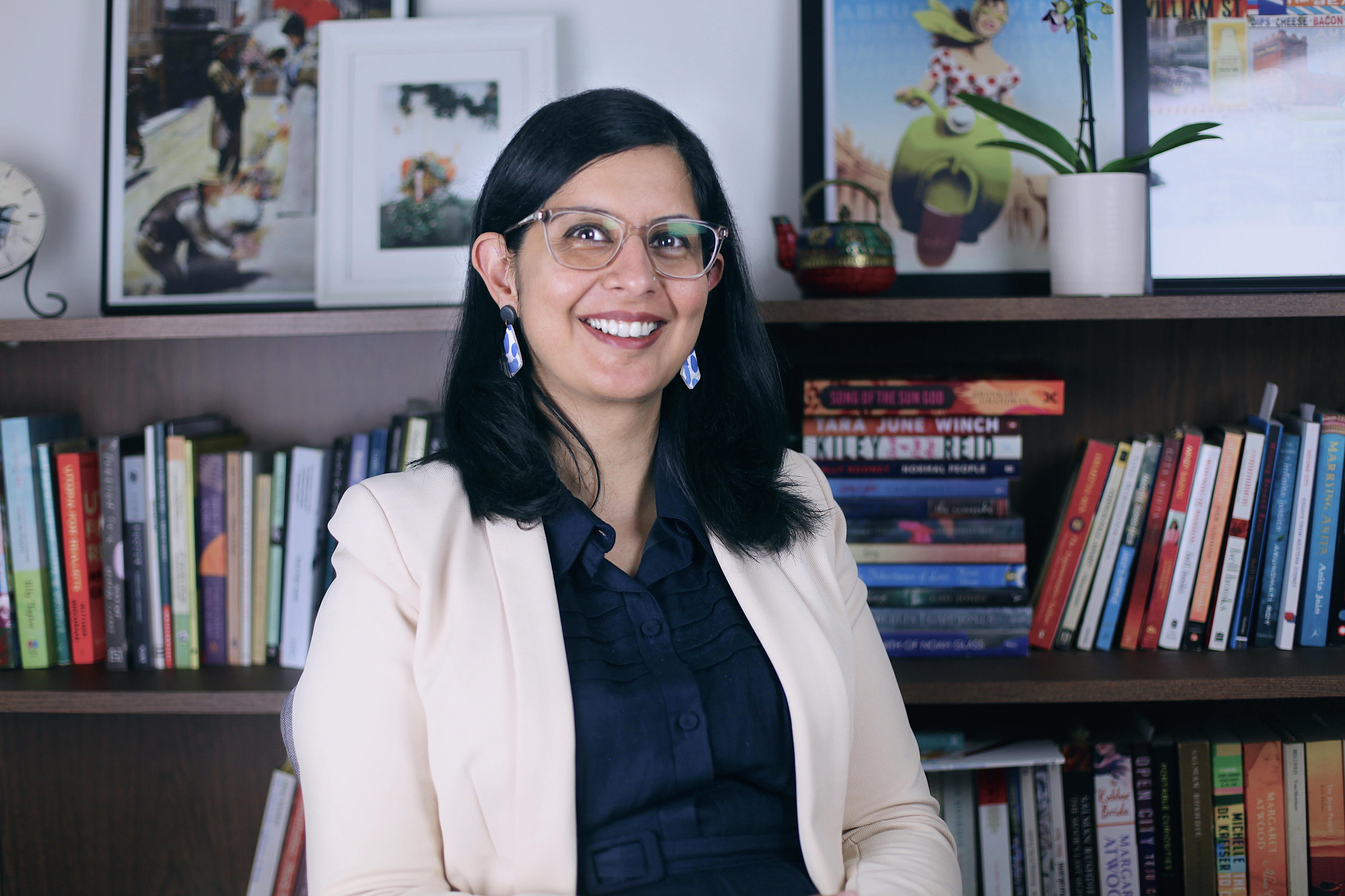 Sukhmani Khorana, with shoulder-length brown hair, glasses and cream jacket, sits smiling in an office before full bookshelves.