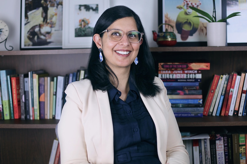 Sukhmani Khorana, with shoulder-length brown hair, glasses and cream jacket, sits smiling in an office before full bookshelves.