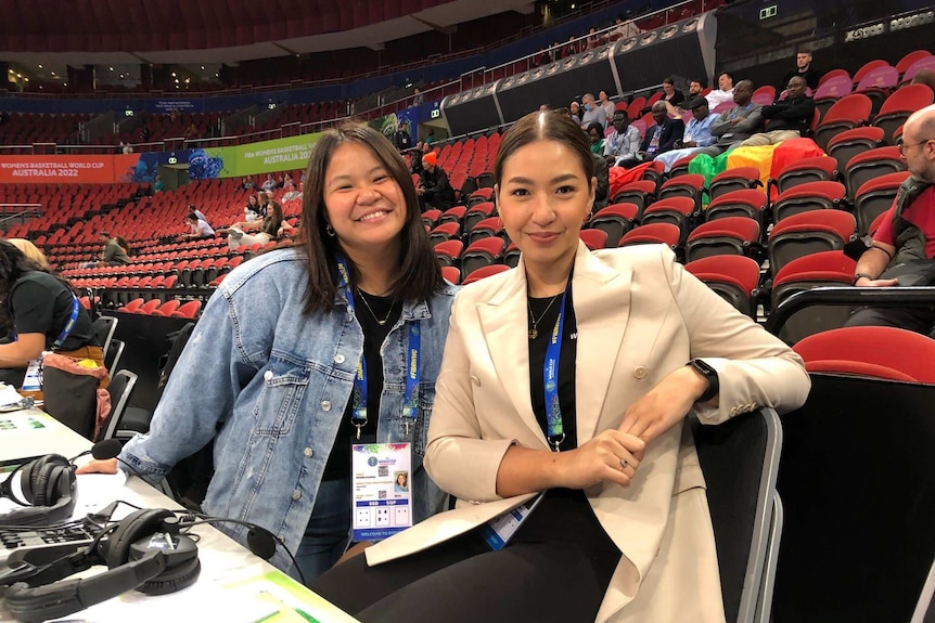 Two women wearing lanyards at a commentators box in a stadium pause to smile to camera.