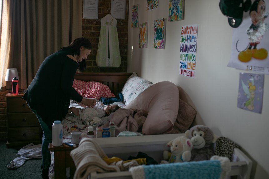 A woman changes the nappy of a baby in a bedroom with a cot and various baby toys.