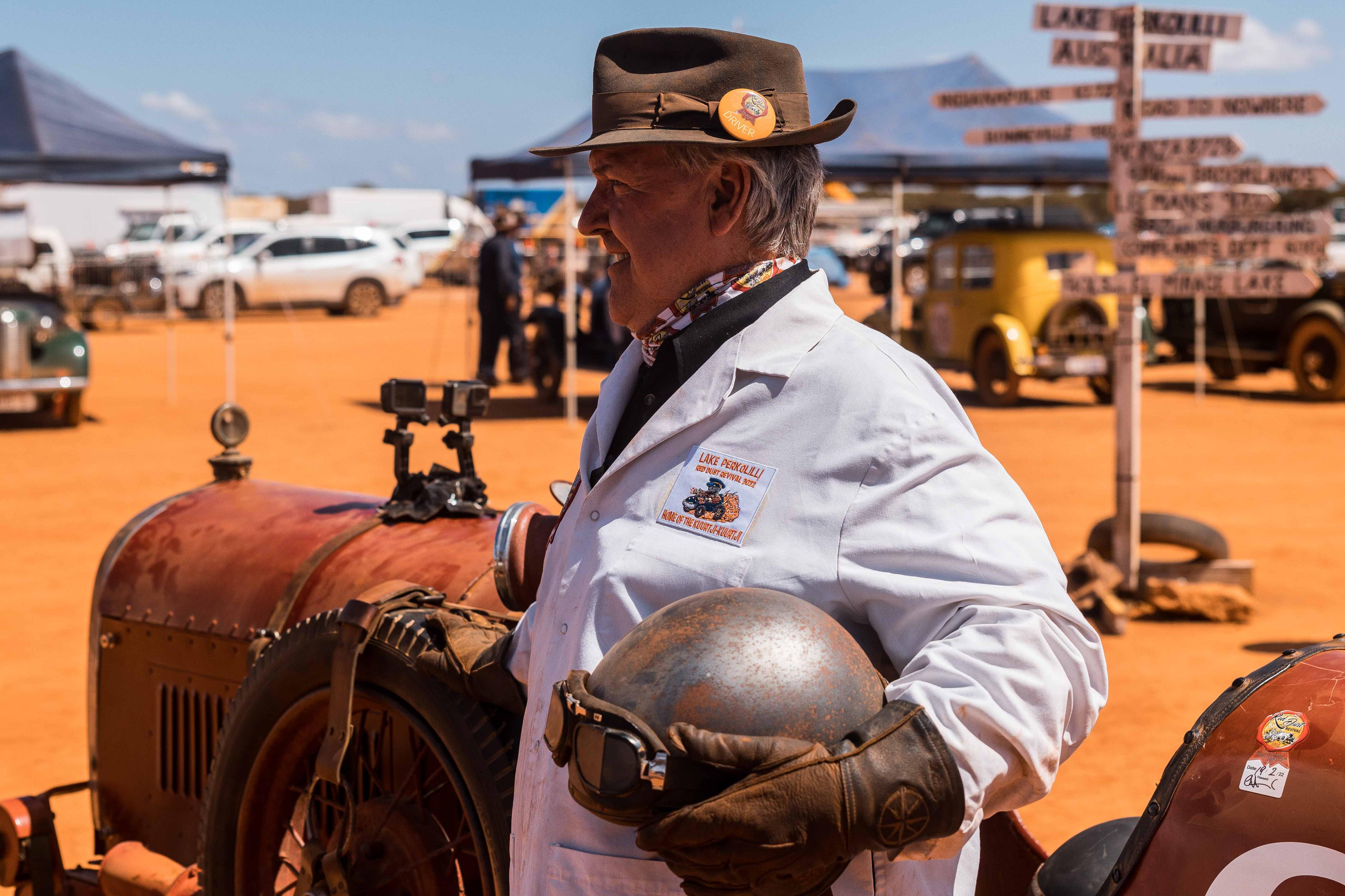 A man standing next to a vintage car holding a helmet and goggles.  